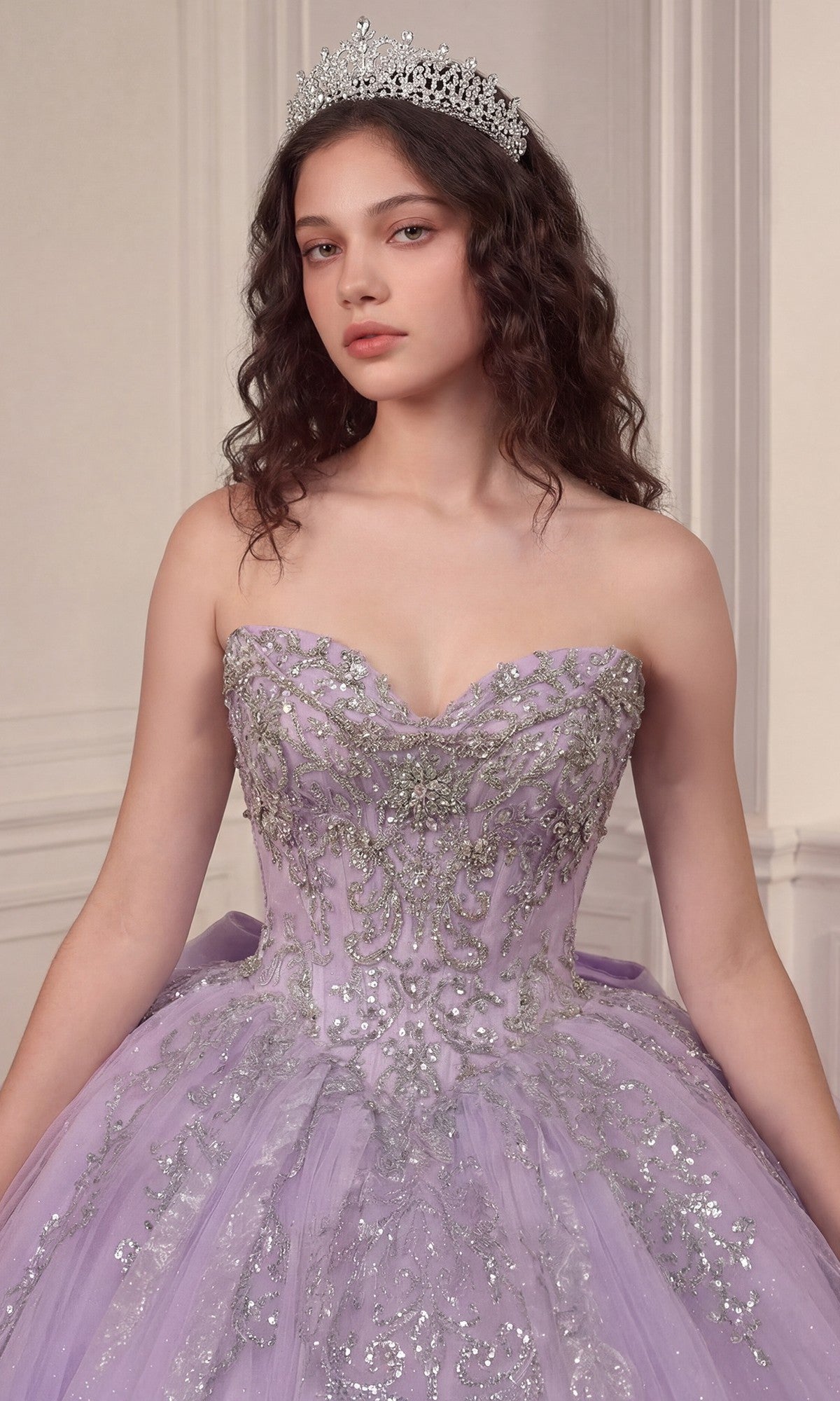 A young woman wearing the Quinceanera Dress 15730 By Ladivine—a lavender ball gown featuring a crystal-embellished corset bodice and a glitter tulle skirt—paired with a silver tiara, stands before an ornate neutral wall, gazing at the camera.